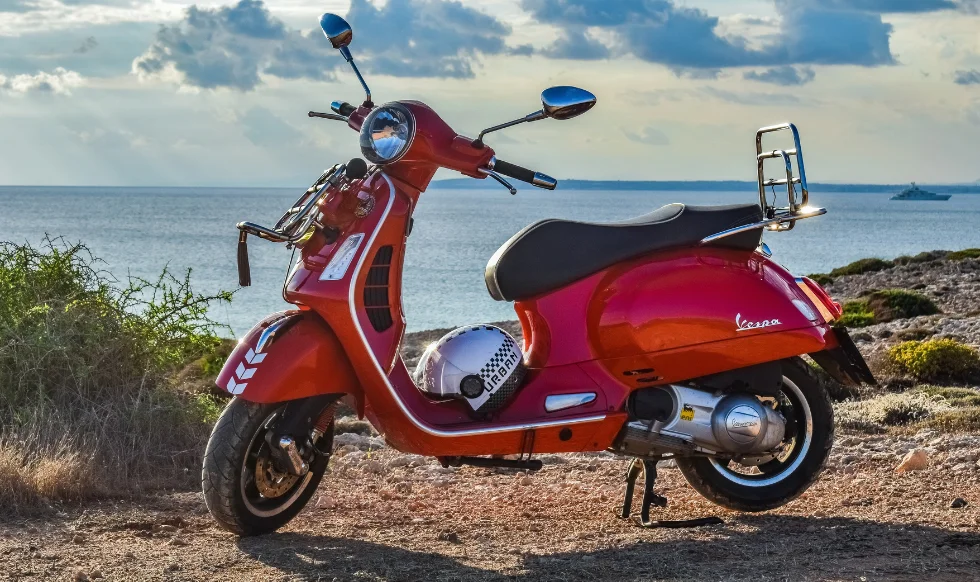 A vibrant red Vespa scooter parked on a rocky coastal path with the ocean and a distant boat in the background.