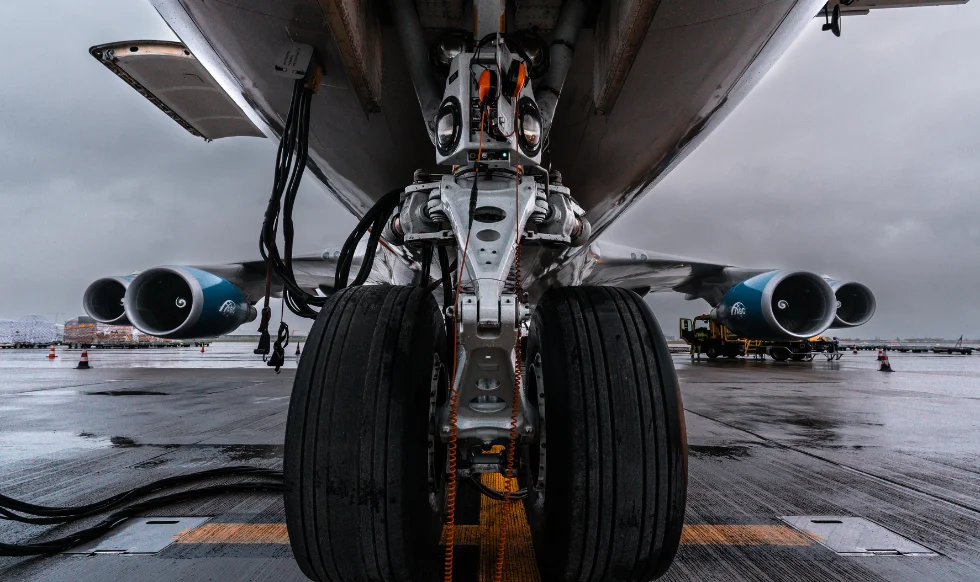Close-up view of an aircraft's landing gear and tires on a wet airport runway, with engines and terminal visible in the background under cloudy skies.