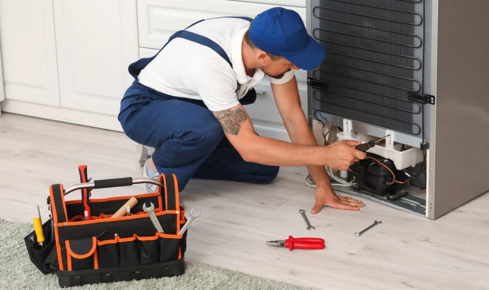 A repair technician in blue overalls and cap using tools to fix the back of a refrigerator, with a toolbox nearby on the floor