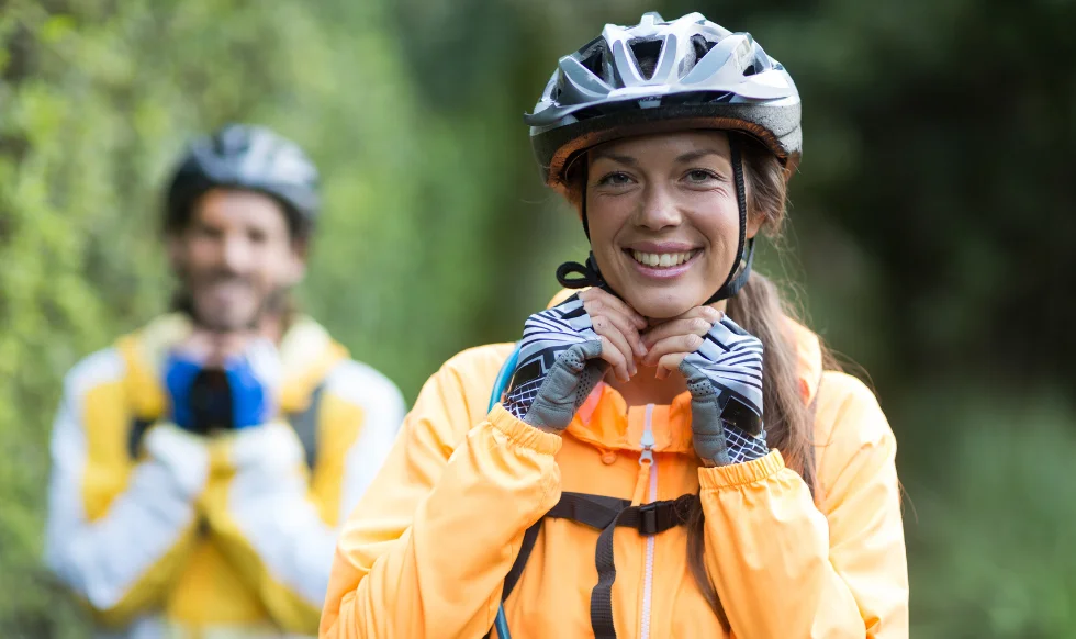 Woman cyclist adjusting her helmet strap while smiling, with another cyclist in the background on a nature trail.