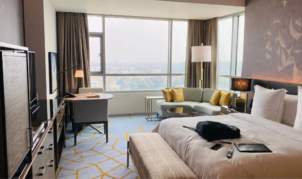 Spacious modern hotel room with large windows, a king-sized bed, a work desk, a corner sofa, and various personal items placed on the bed.