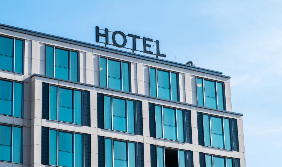 A modern hotel building with large glass windows and a prominent "HOTEL" sign on the rooftop against a clear blue sky.
