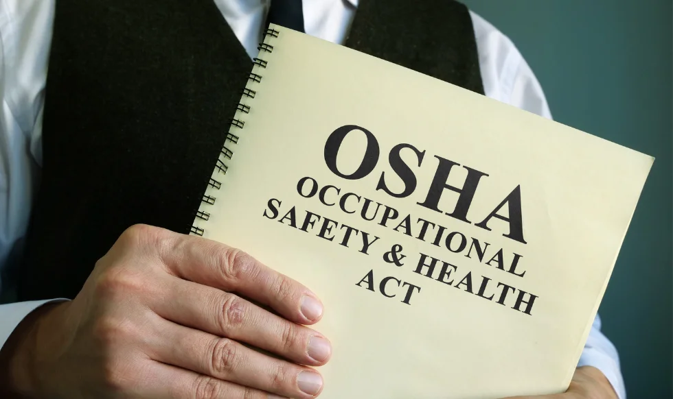 A person holding a spiral-bound booklet titled "OSHA Occupational Safety & Health Act," representing workplace safety regulations and legal compliance.