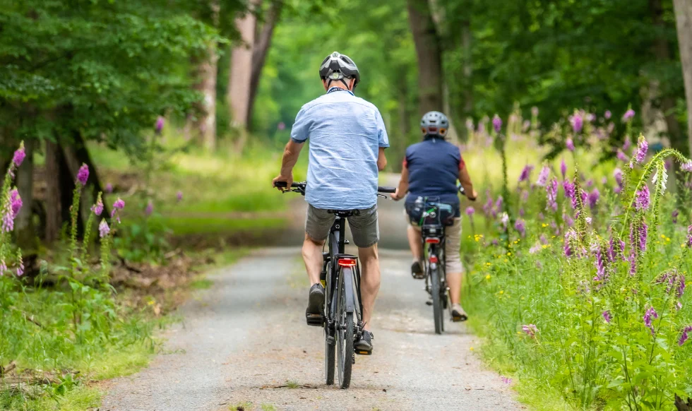 Two people wearing helmets cycling on a forest path surrounded by greenery and wildflowers