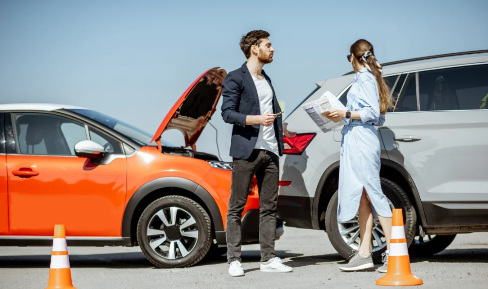 Man and woman discussing after a car accident, with orange car and gray SUV damaged, standing on road with traffic cones