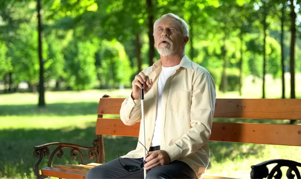 An elderly blind man with a white cane sitting on a park bench, looking contemplative in a sunlit, green park.