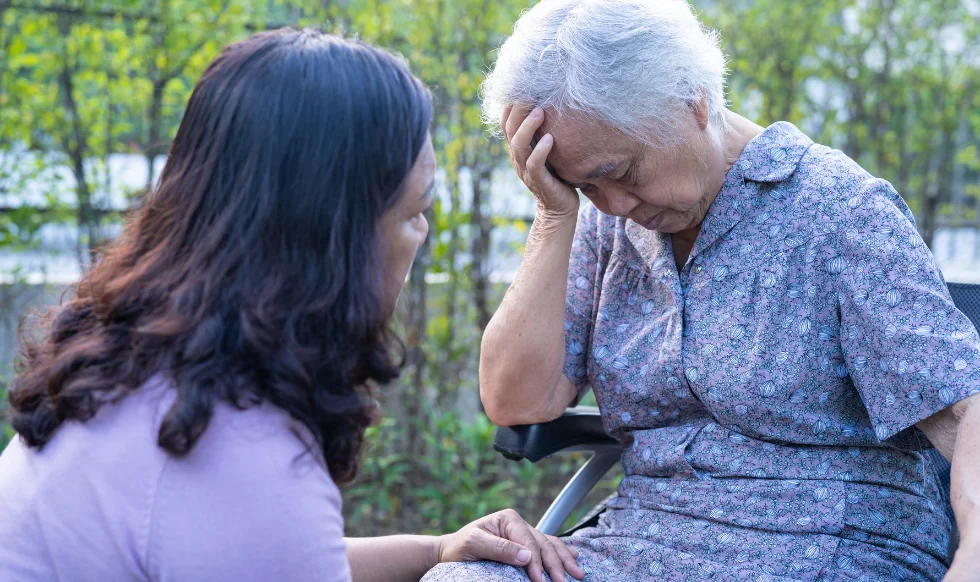 An elderly woman with white hair looks distressed while sitting outside in a wheelchair, holding her head. A younger woman, possibly a caregiver or family member, offers support by gently touching her knee.