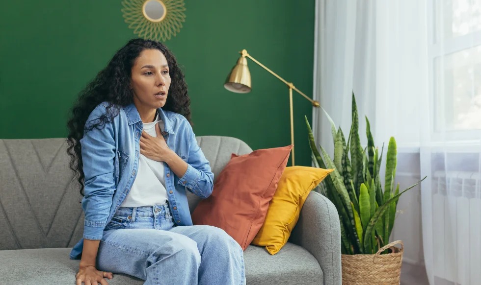 A distressed woman sitting on a couch holding her chest, showing signs of anxiety or a panic attack, possibly related to emotional trauma.