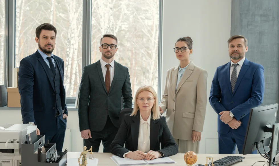 Five professional personal injury lawyers in formal business attire standing and sitting confidently in a modern office