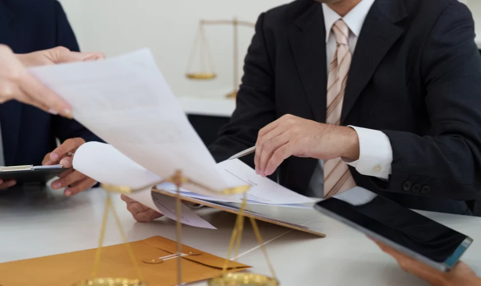 A group of lawyers in formal attire reviewing legal documents together at a desk with a golden scale of justice in the foreground.